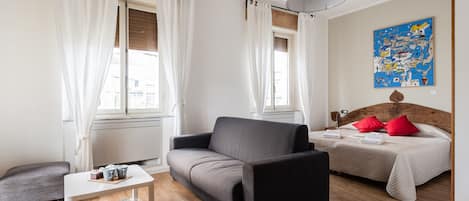 Moderni hotellihuone, jossa on sohva, pieni pöytä kirjoineen, punaisilla tyynyillä varustettu sänky ja suuri, abstrakti taulu seinällä.