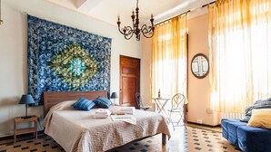 A bedroom with a bed, a chandelier, a tapestry, a wooden door, a small table, and a blue sofa. - La Gemma di Elena (Lucca)