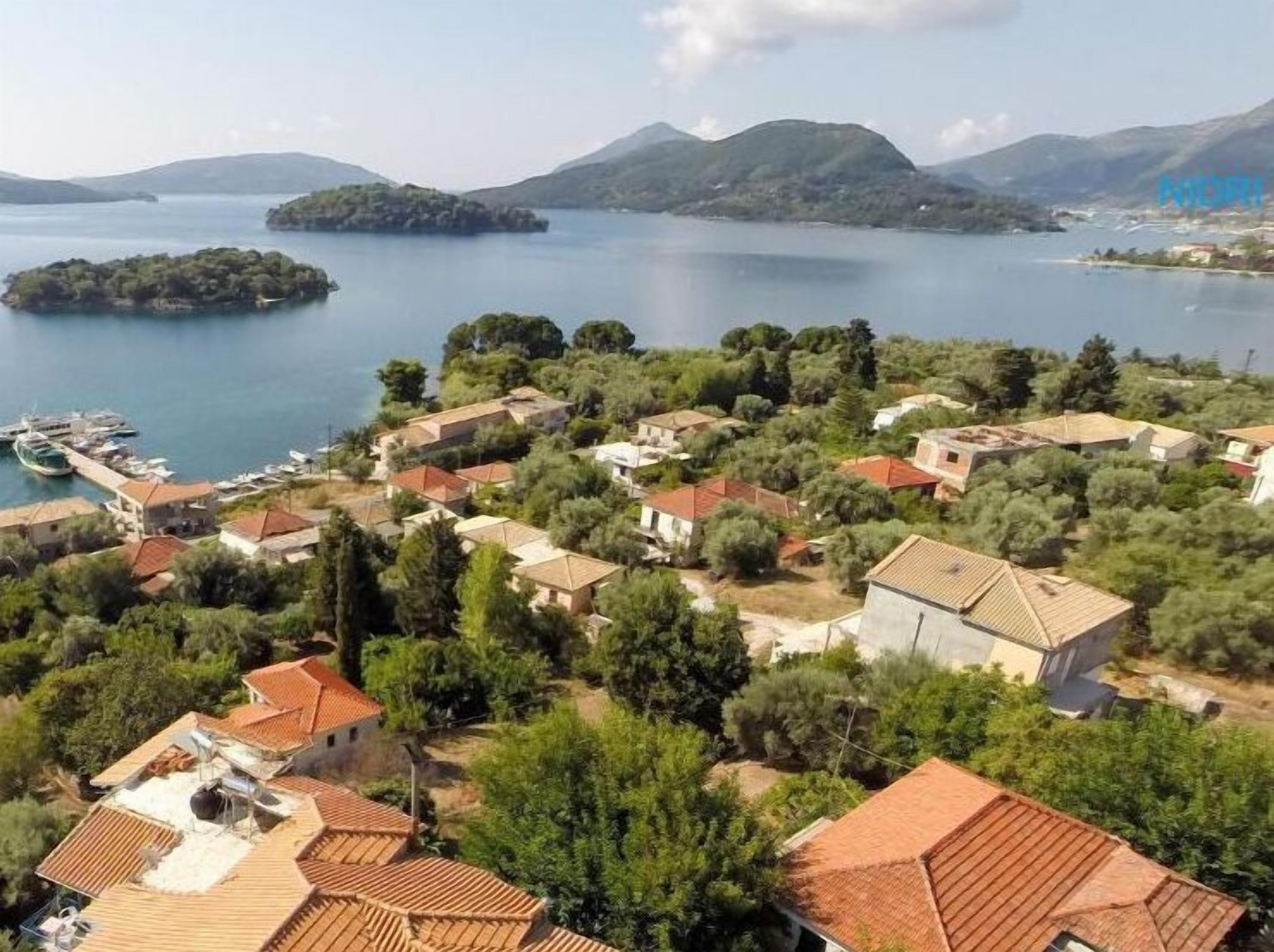 A coastal village with terracotta-roofed houses, a marina, and a backdrop of mountains and a lake.