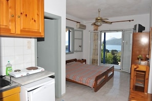 A bedroom with a bed, a ceiling fan, a balcony with a view, and a kitchen area with cabinets and a stove. - Gianna Studios (Lefkada)