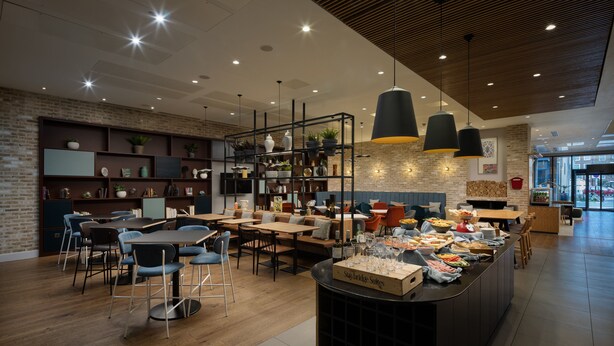 A modern cafe with wooden tables and chairs, a central buffet area, and shelves with plants and books. - Staybridge Suites London - Vauxhall by IHG (London)