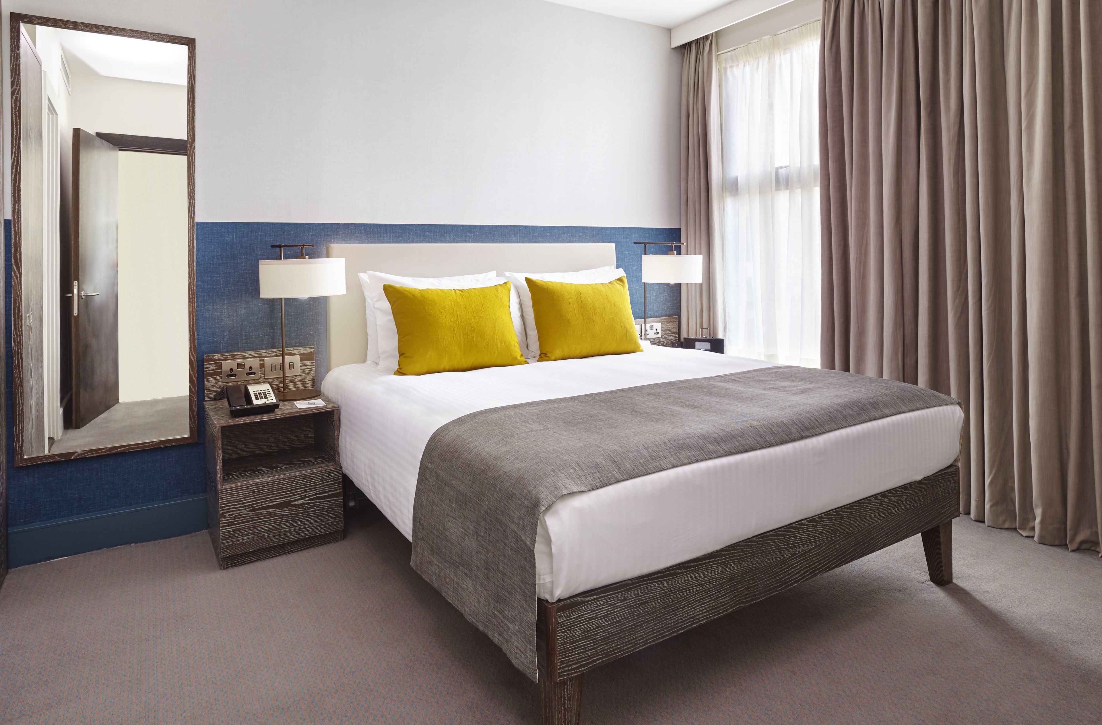 A hotel room with a large bed, two bedside lamps, a nightstand, a mirror, and a window with curtains.