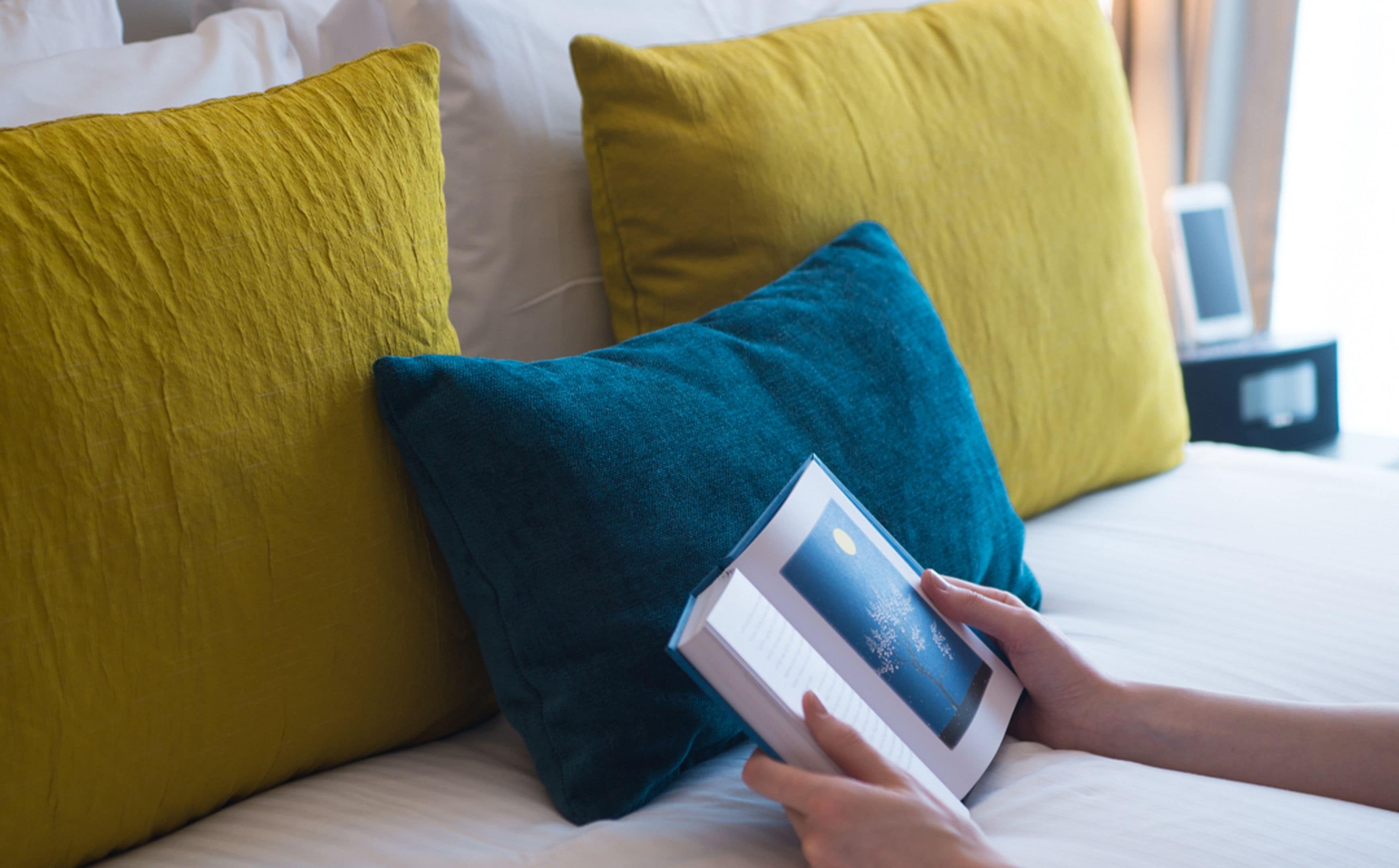 A person reading a book on a bed with yellow and teal pillows.