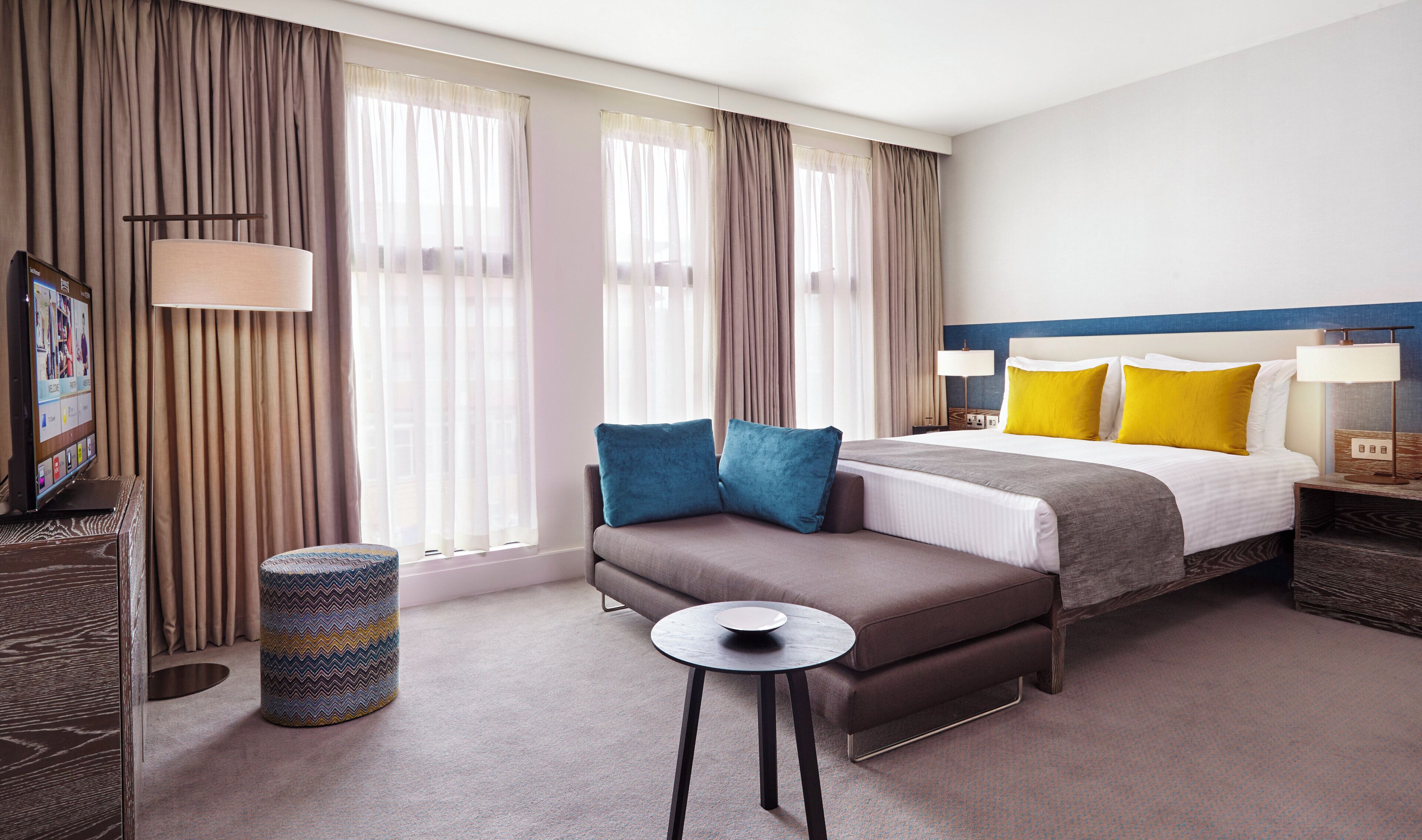 A hotel room with a bed, bedside tables, a sofa, a small round table, and curtains.