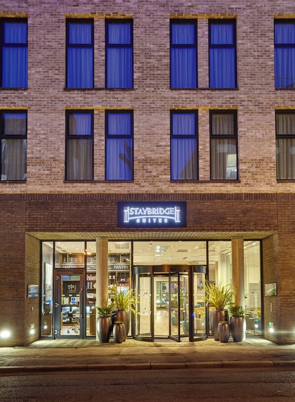 Entrada de un hotel Staybridge Suites con fachada de ladrillo, plantas en macetas grandes y puerta de acceso de vidrio.