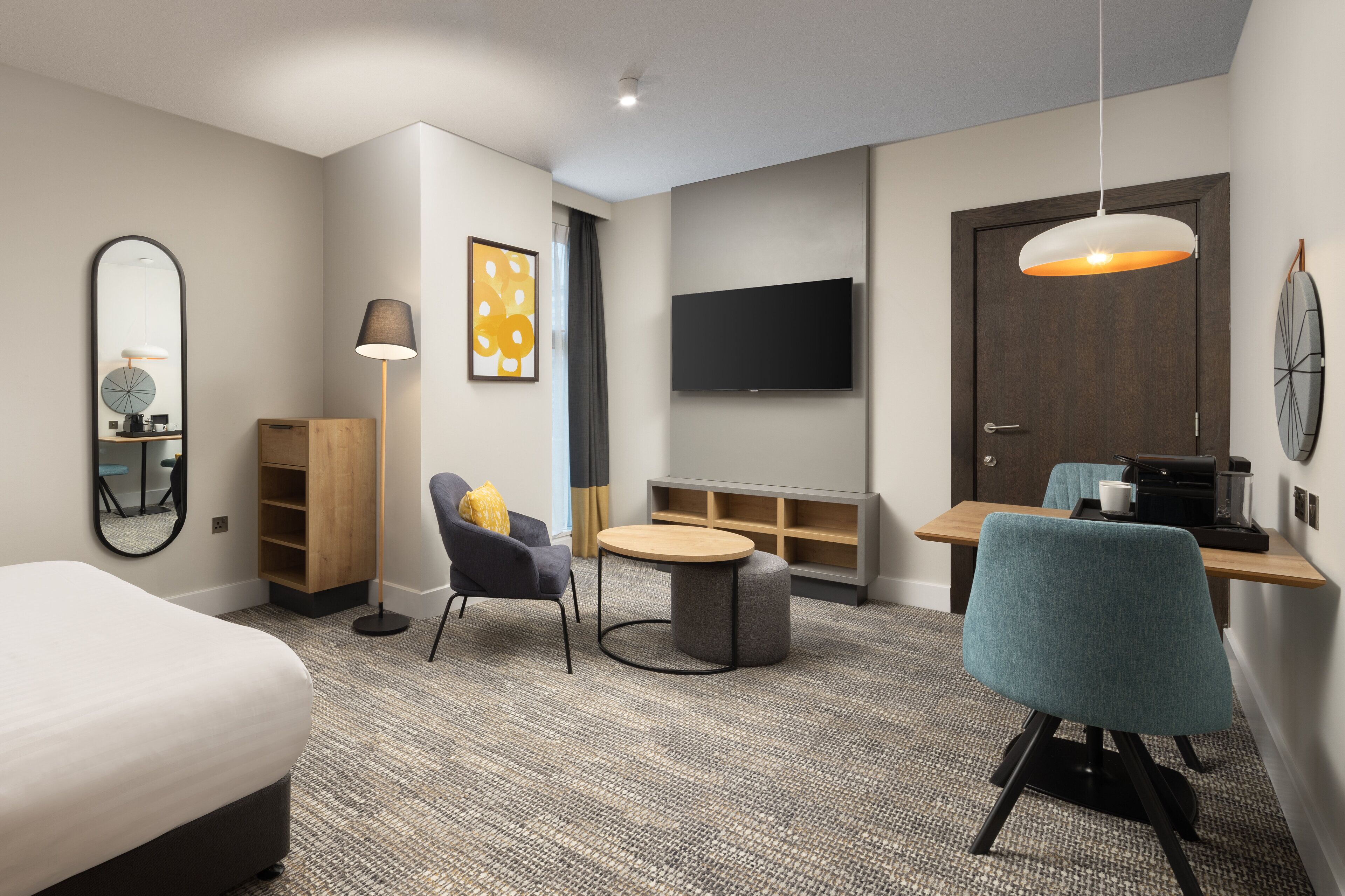 A modern hotel room with a bed, a round table, two chairs, a TV, a bookshelf, and a desk with a lamp.