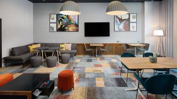 A modern hotel room with a sectional sofa, a small table, a dining table, and a flat-screen TV mounted on the wall.