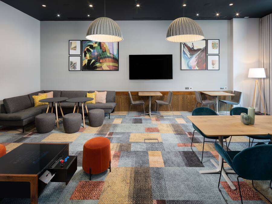 A modern hotel room with a sectional sofa, a small table, a dining table, and a flat-screen TV mounted on the wall.