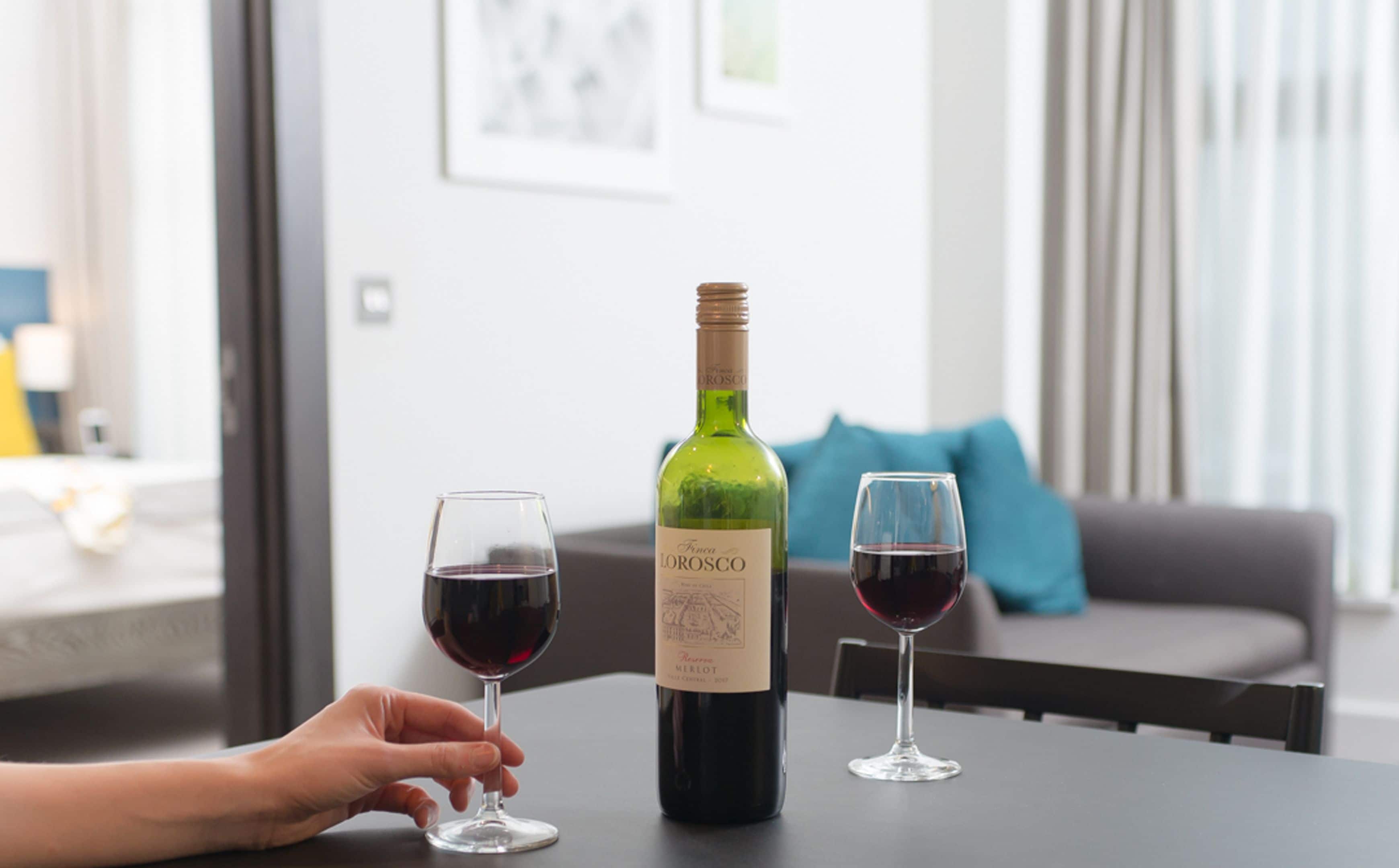 A bottle of wine and two glasses on a table in a modern living room with a grey sofa and a blue pillow in the background.