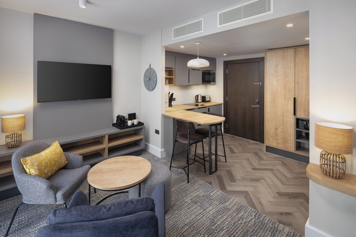 A modern living room with a flat-screen TV, grey sofas, a round coffee table, and a herringbone-patterned floor.