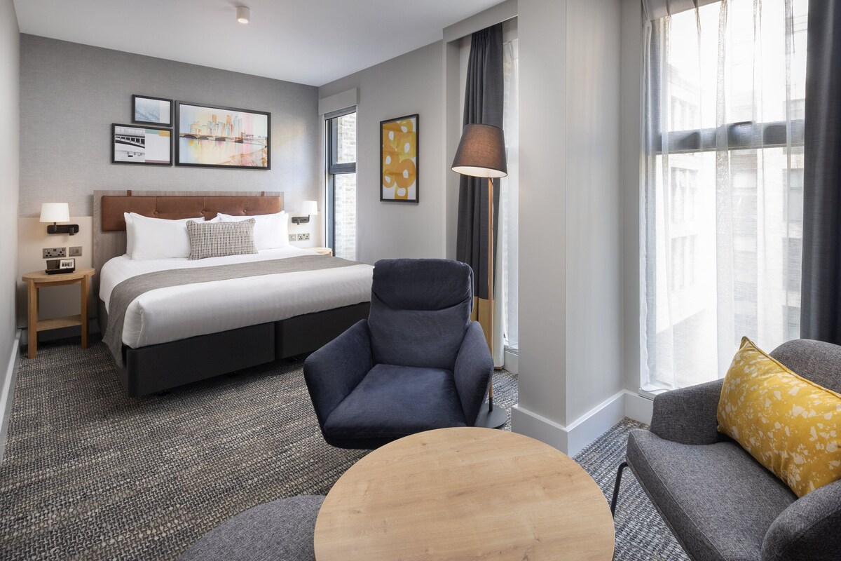 A hotel room with a large bed, a chair, a sofa, and a small table. There are framed pictures on the wall and a lamp next to the bed.