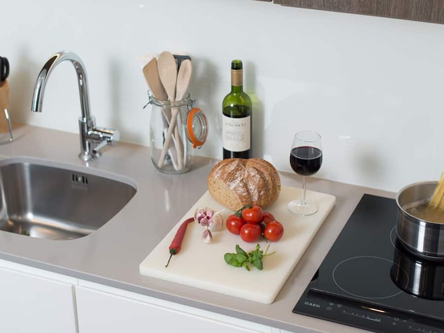 A kitchen with a sink, a cutting board with tomatoes, garlic, and a loaf of bread, a bottle of wine, and a pot of spaghetti on an induction cooktop.