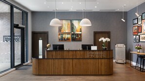 A reception desk with a wooden finish, two pendant lights, and a cityscape painting above. - Staybridge Suites London - Vauxhall by IHG (London)