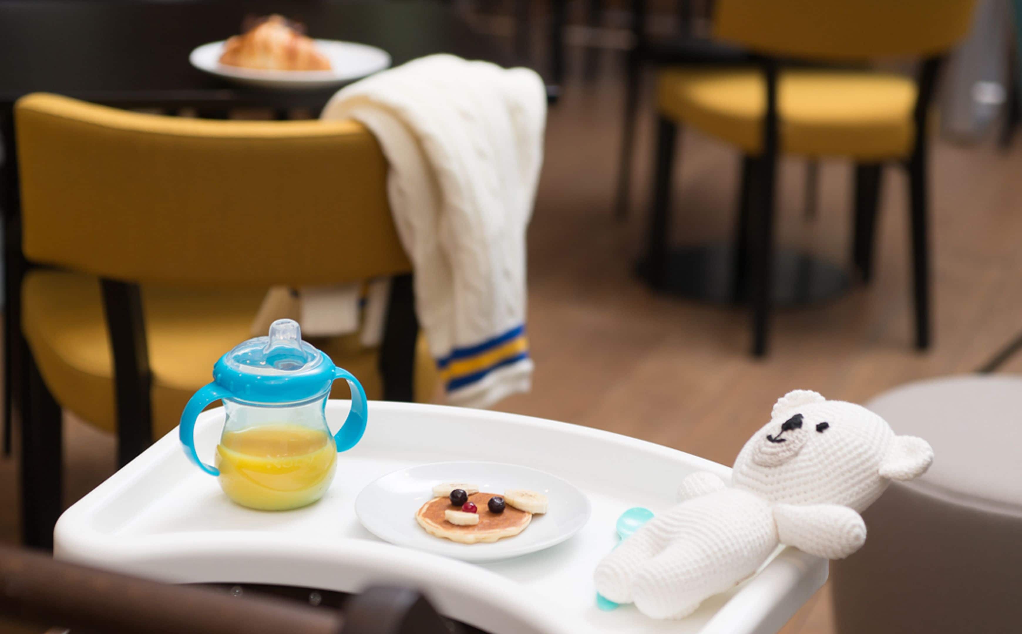 A dining table with a baby high chair, a baby bottle, a plate with pancakes, and a teddy bear.