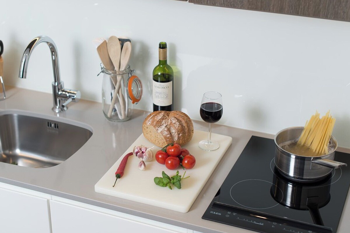 A kitchen counter with a sink, a bottle of wine, a glass of red wine, a loaf of bread, tomatoes, garlic, and a pot of spaghetti.