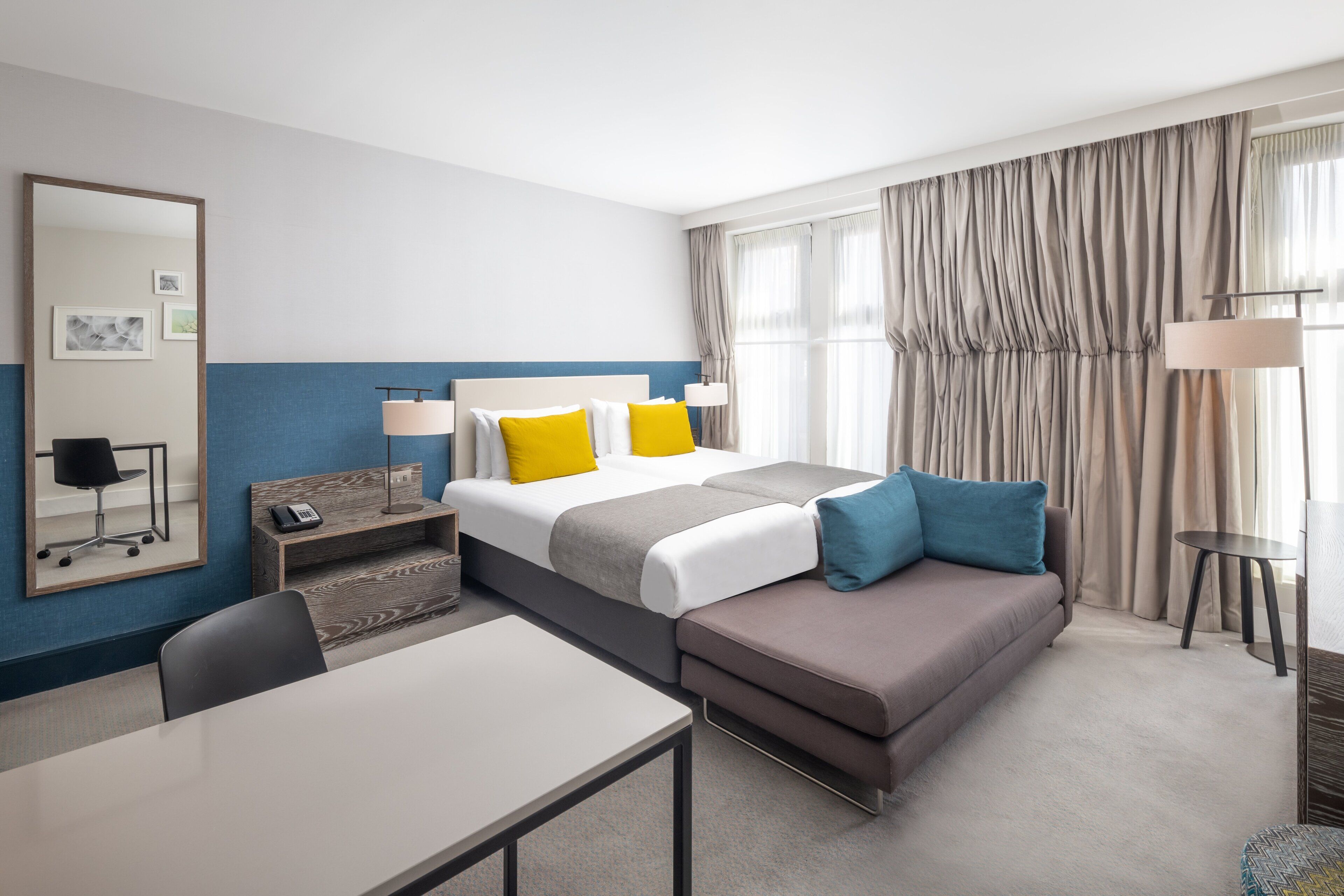 A hotel room with a bed, a sofa, a desk, a chair, a lamp, and a mirror.