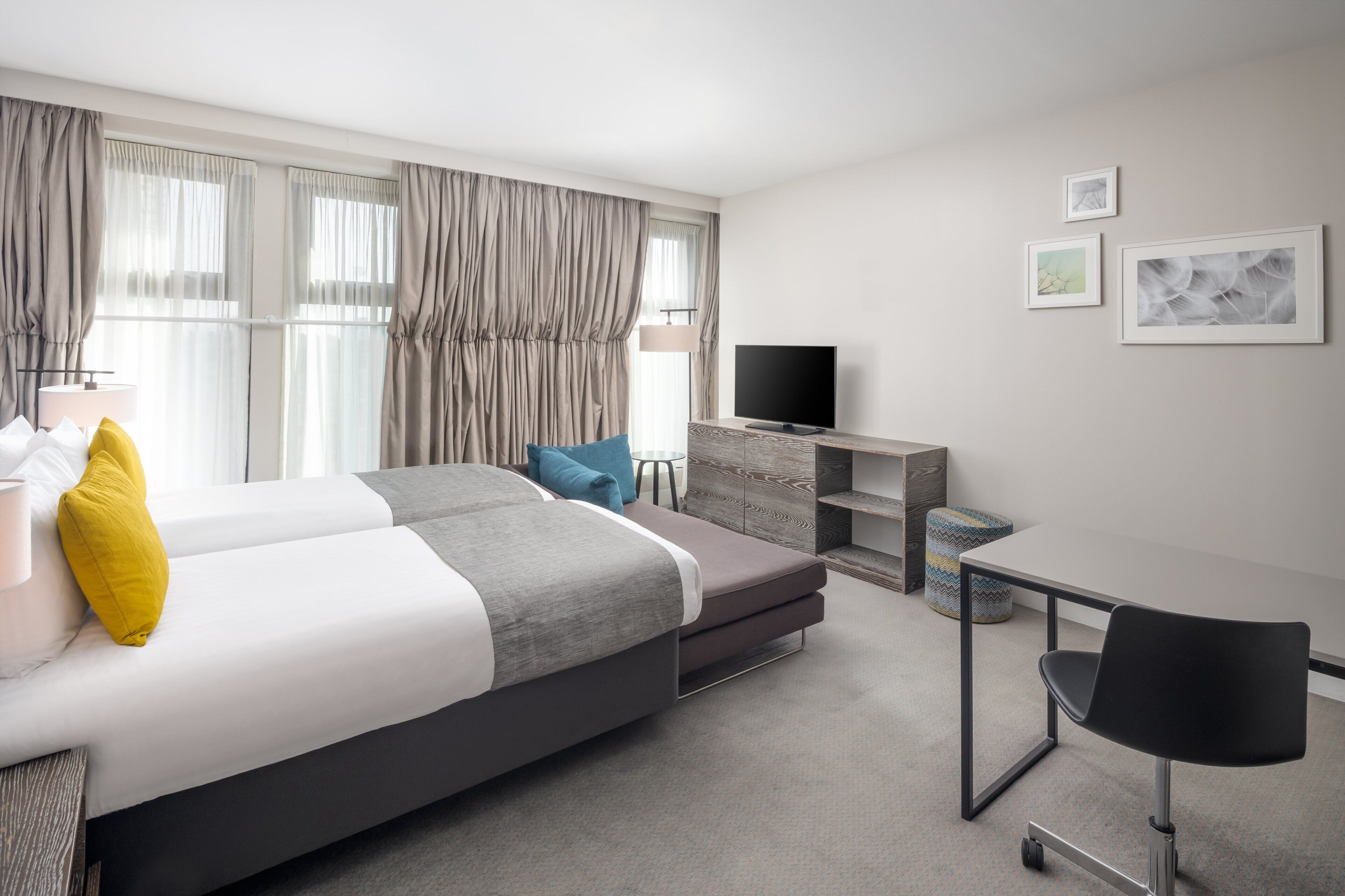 A modern hotel room with a large bed, a desk, a chair, a television, and artwork on the wall.