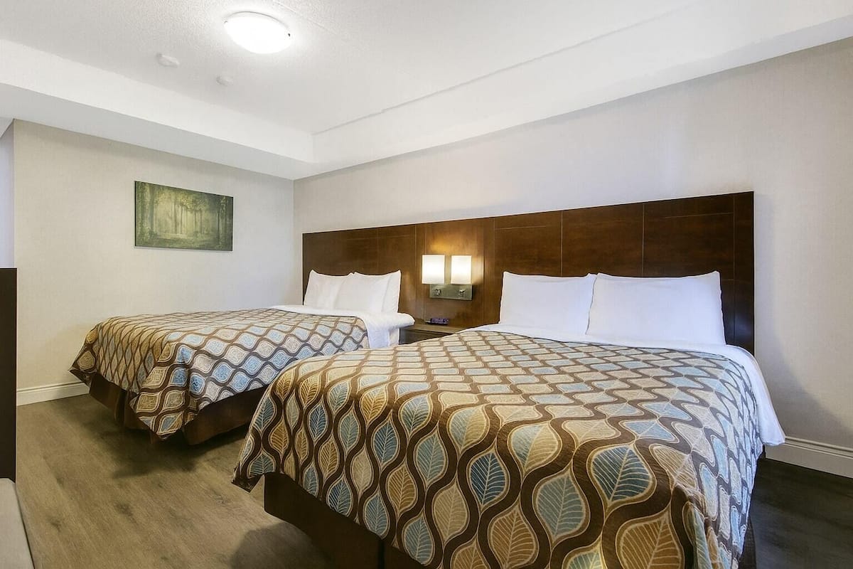 A hotel room with two beds, each with patterned bedding and a wooden headboard. There is a wall-mounted night light and a painting on the wall.