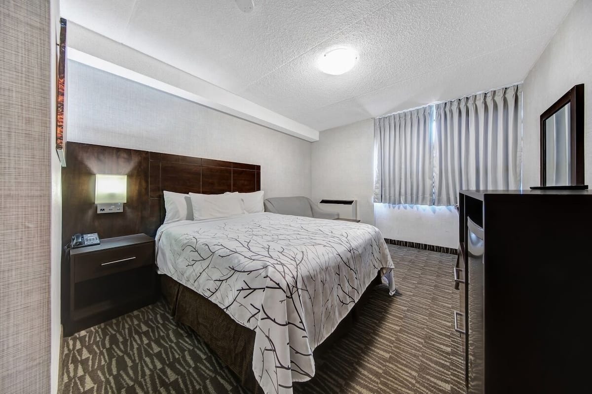 A hotel room with a bed, a bedside table with a phone, a desk with a computer, a chair, and a large window with curtains.