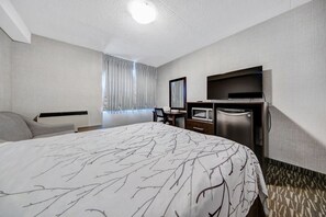 A hotel room with a large bed, a desk with a flat-screen TV, a minibar, and a window with curtains. - Deluxe Inn (Toronto)