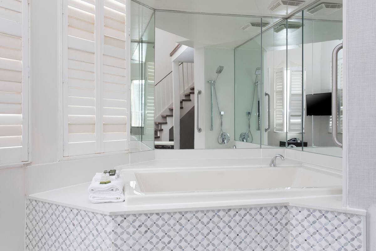 Un baño moderno con una bañera blanca grande, cabina de ducha de vidrio y una escalera visible al fondo.