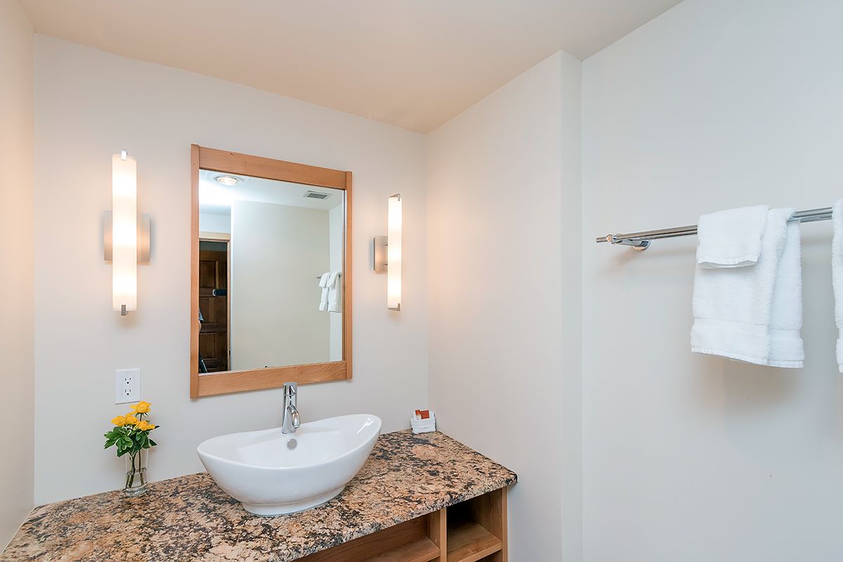 A bathroom with a white sink, granite countertop, a mirror with a wooden frame, two wall-mounted lights, and a towel rack with white towels.