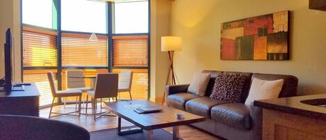 A modern living room with a leather sofa, a coffee table, and a desk with chairs. There is a large window with blinds, a floor lamp, and a painting on the wall.