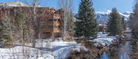 Un chalet au revêtement extérieur en bois, une rivière et, au loin, des montagnes enneigées.