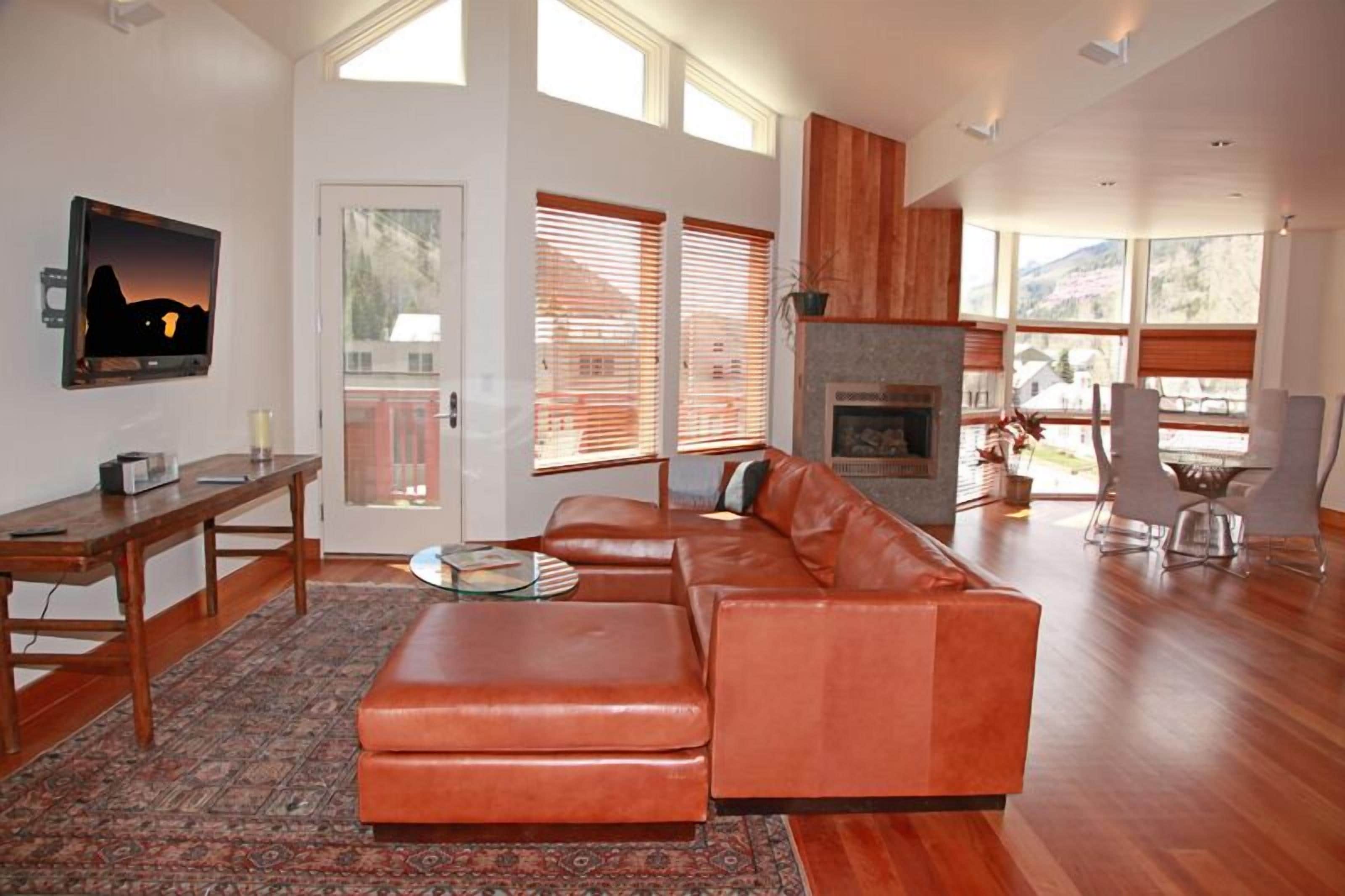 A modern living room with a large brown leather sofa, a glass coffee table, and a fireplace. There is a TV mounted on the wall and a dining area with a table and chairs.