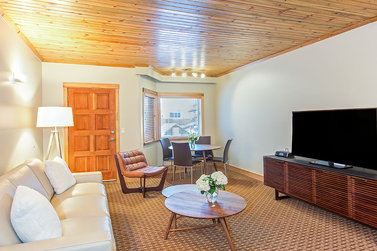 A living room with a wooden ceiling, a sofa, a TV, a dining table, and a window with blinds.