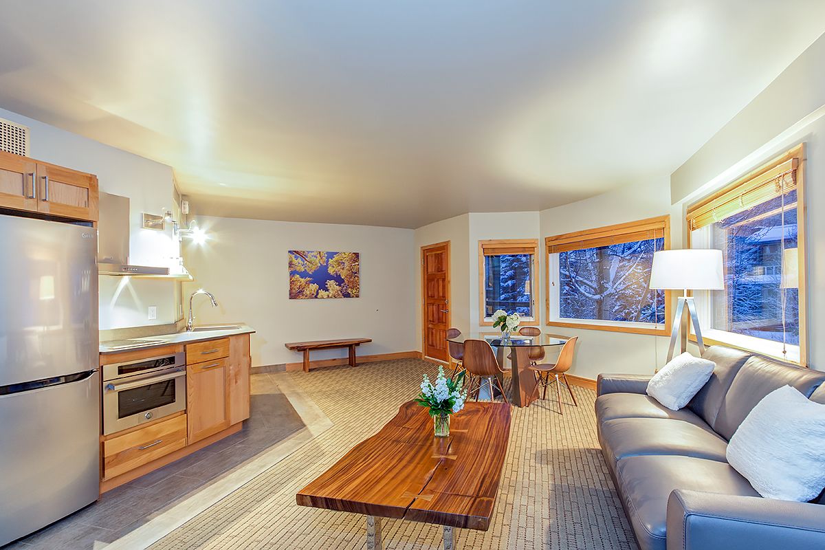 A modern kitchen with built-in appliances, a dining area with a wooden table and chairs, a sofa, and large windows with blinds.