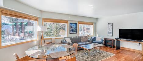 A spacious living room with a glass dining table, comfortable seating, a fireplace, and large windows offering a view of the outside.