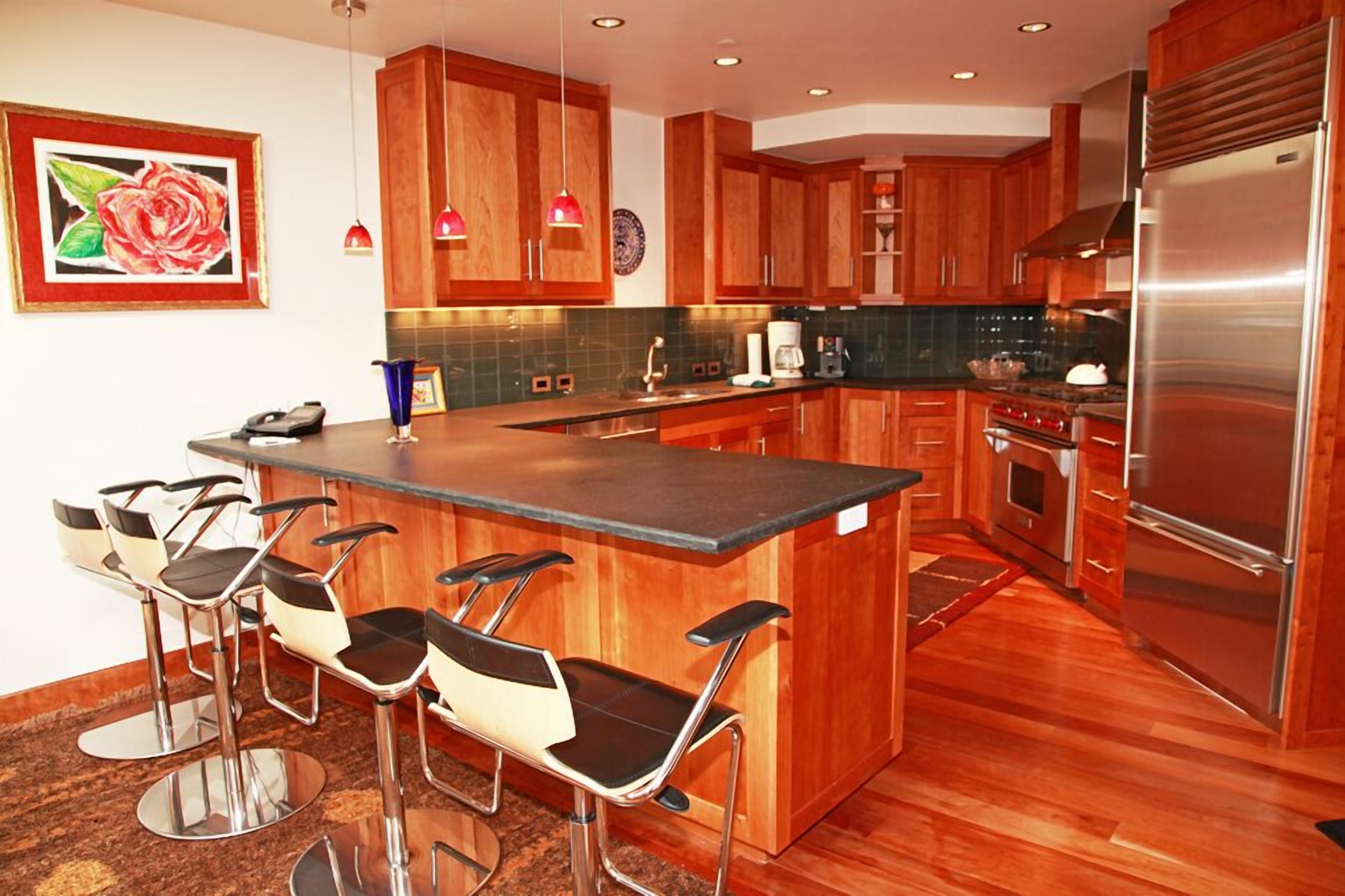 A modern kitchen with wooden cabinets, a stainless steel refrigerator, and a bar area with stools.