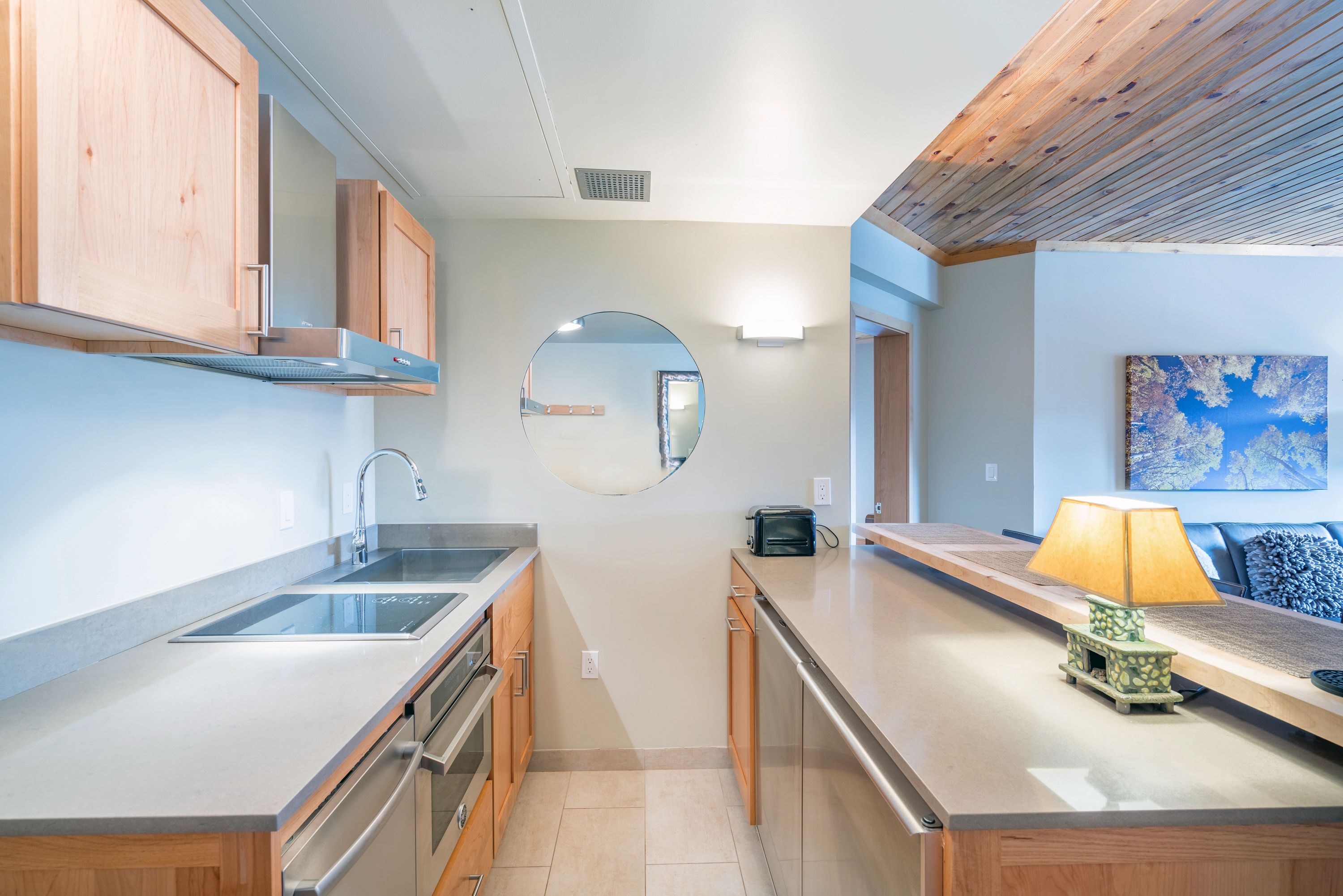 Una cocina moderna con armarios de madera, electrodomésticos de acero inoxidable, un espejo redondo y una pequeña mesa con lámpara.