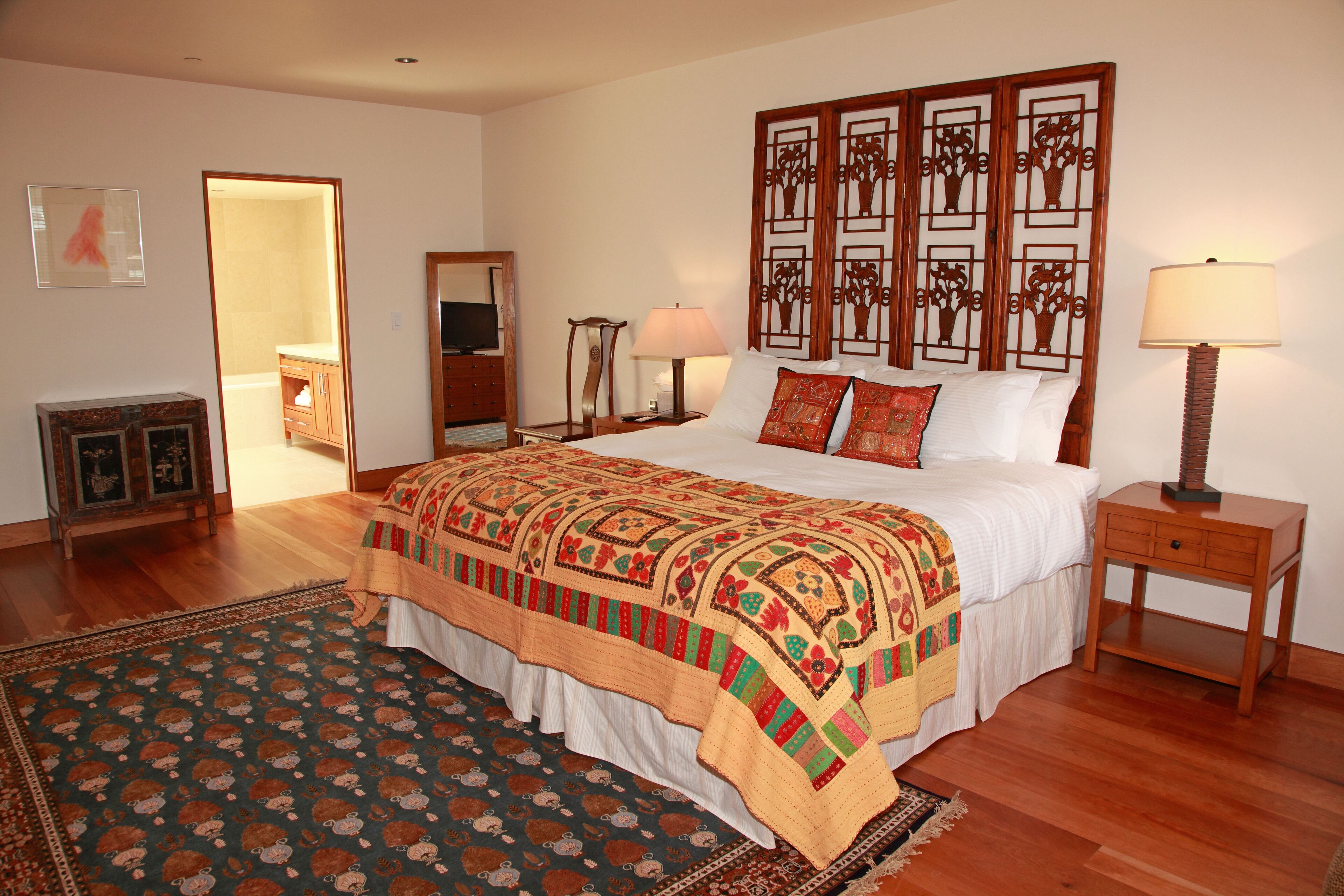 A bedroom with a large bed, a wooden headboard, a bedside table with a lamp, a small cabinet, and a patterned rug.