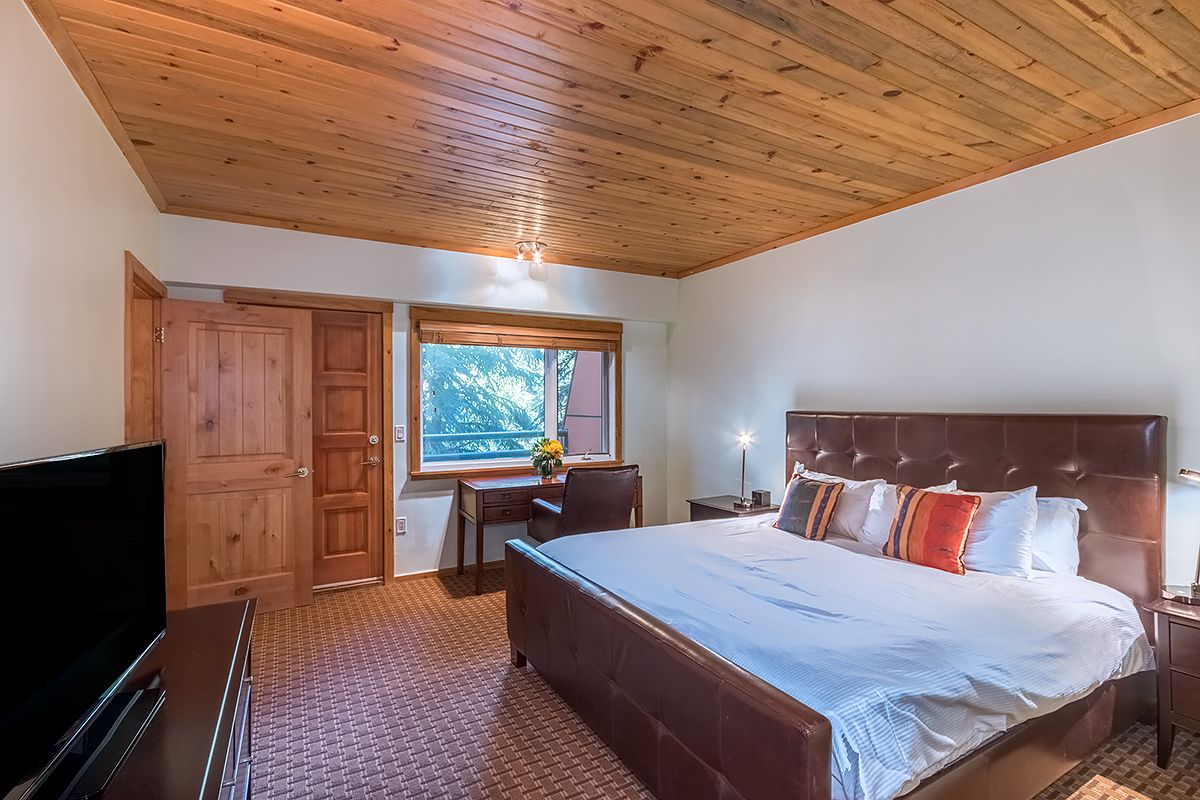 A bedroom with a large bed, wooden ceiling, a TV, a desk with a chair, and a window with a view of trees.
