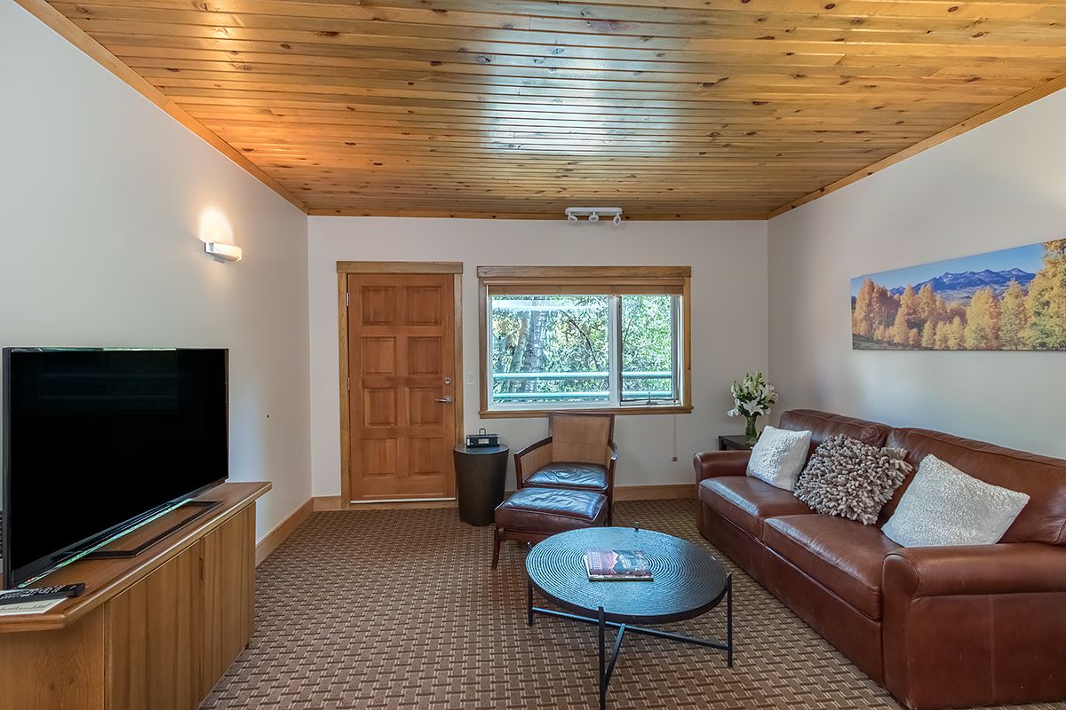A living room with a large flat-screen TV, a brown leather sofa, a round coffee table, a wooden ceiling, and a window with a view of trees.