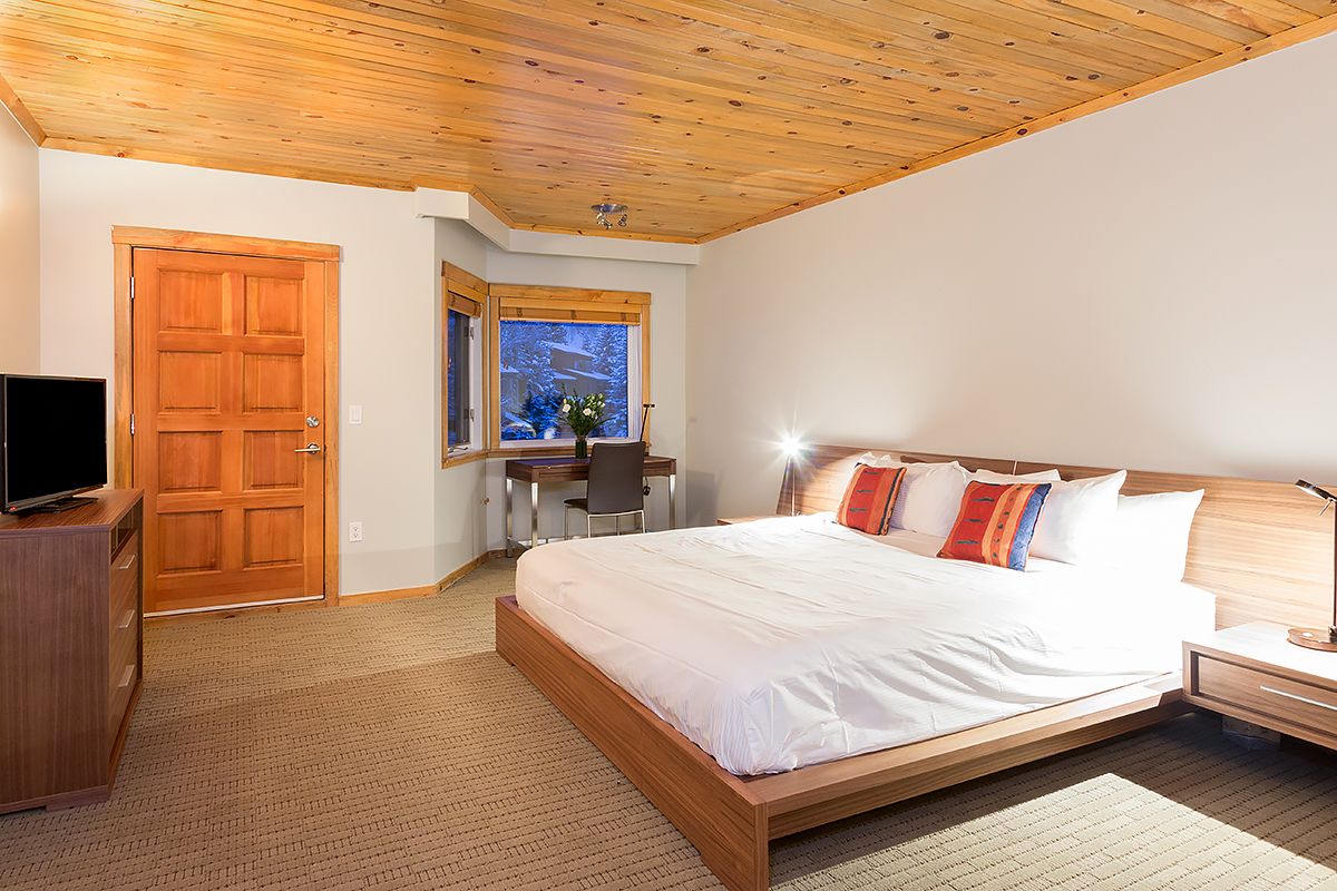 A bedroom with a wooden ceiling, a large bed, a desk, and a TV.