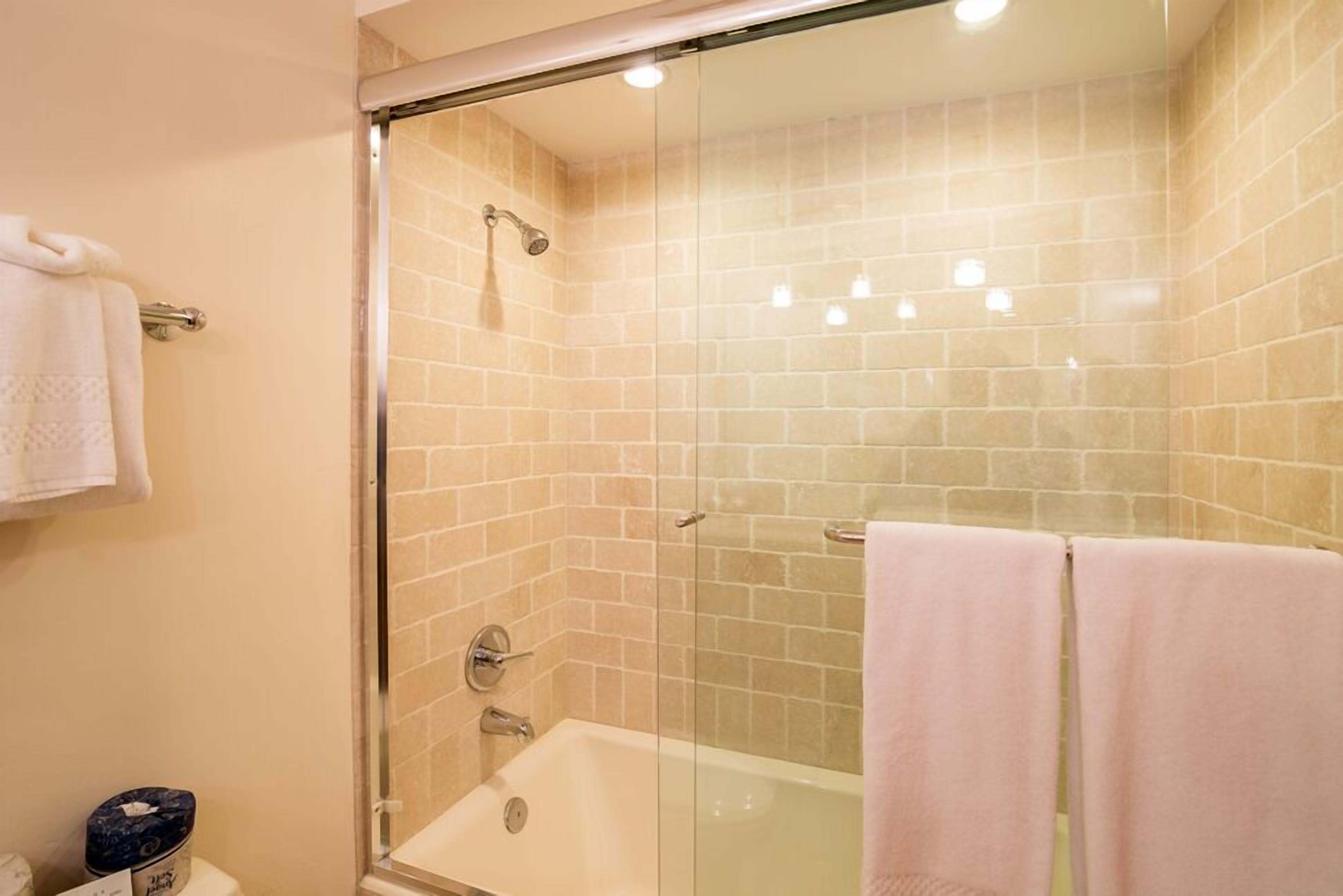 A glass-enclosed shower area with a bathtub, beige tiled walls, and two towels on the floor.