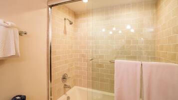 A glass-enclosed shower area with a bathtub, beige tiled walls, and two towels on the floor.