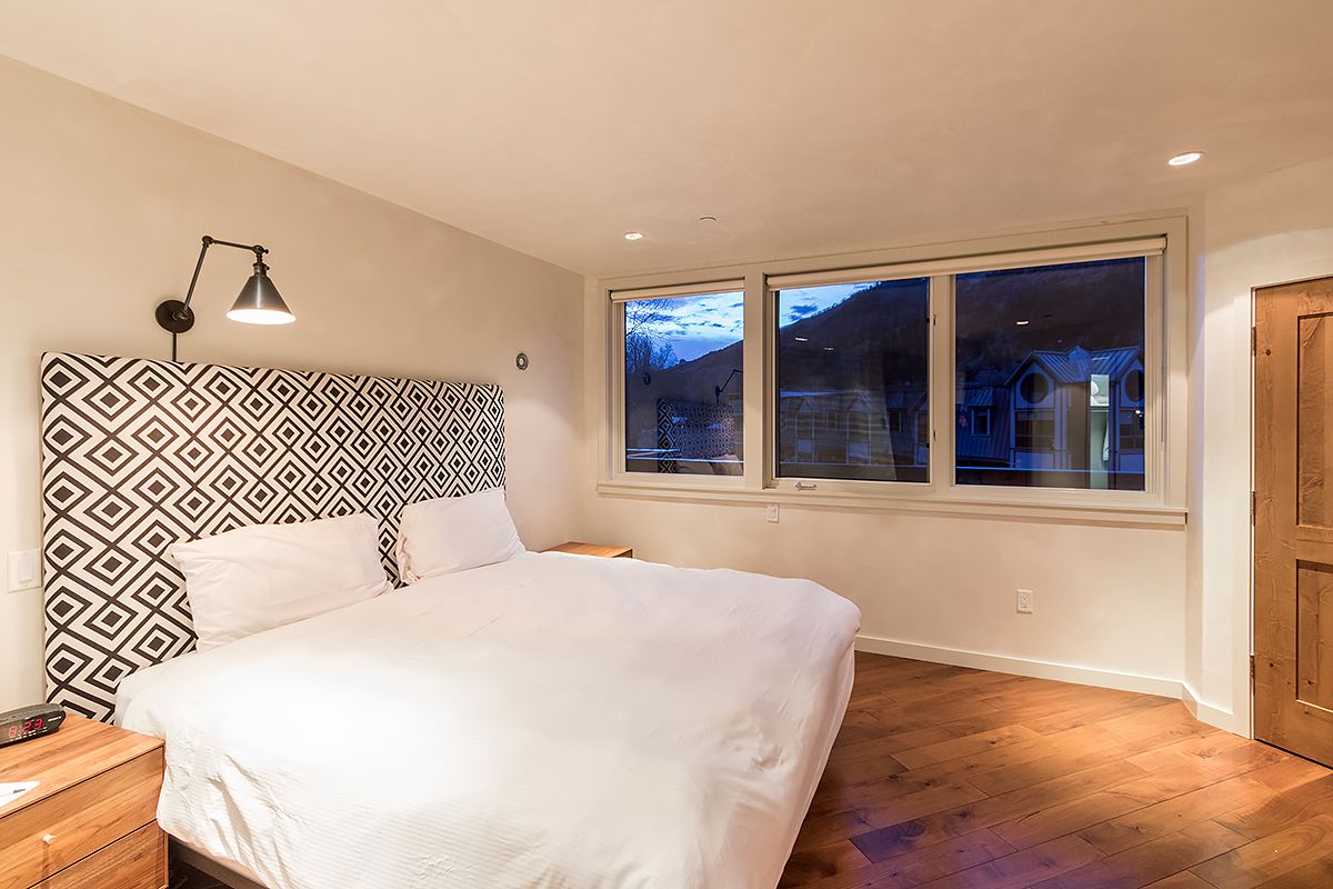 Une chambre à coucher avec une tête de lit à motifs, une grande fenêtre donnant sur des immeubles et un plancher en bois.