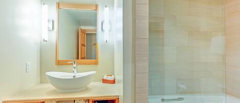 A modern bathroom with a large white sink, a wooden vanity, a glass shower enclosure, and a mirror above the sink.