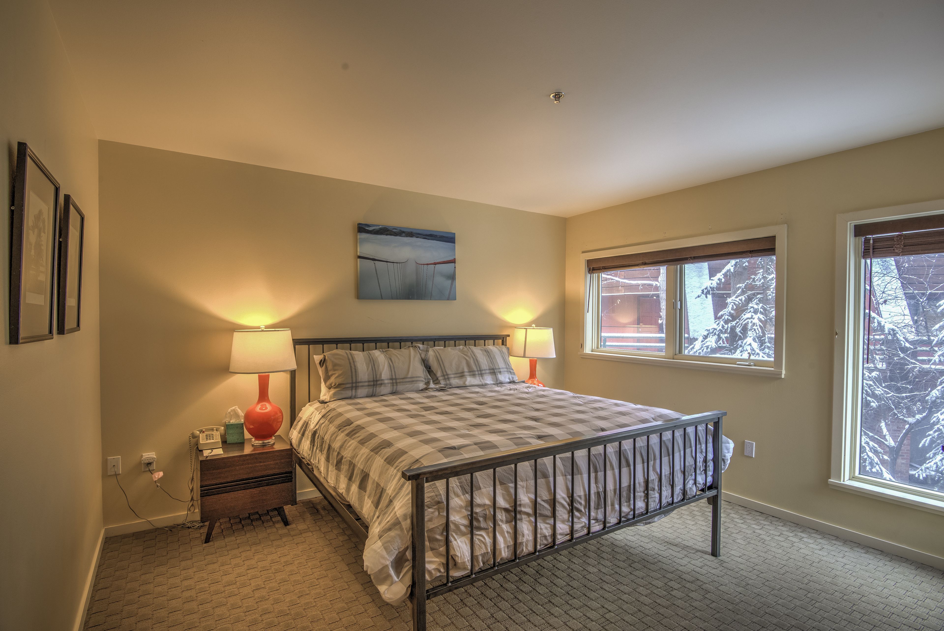A bedroom with a bed, two bedside lamps, a nightstand, a wall-mounted painting, and a window with a view of trees.