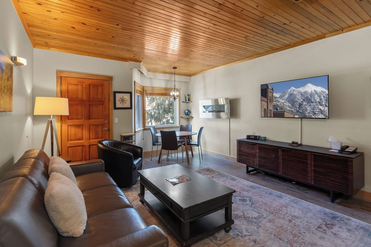 Una sala de estar con techo de madera, un sofá de cuero marrón, una mesa de centro, un televisor y un comedor con sillas y una mesa.