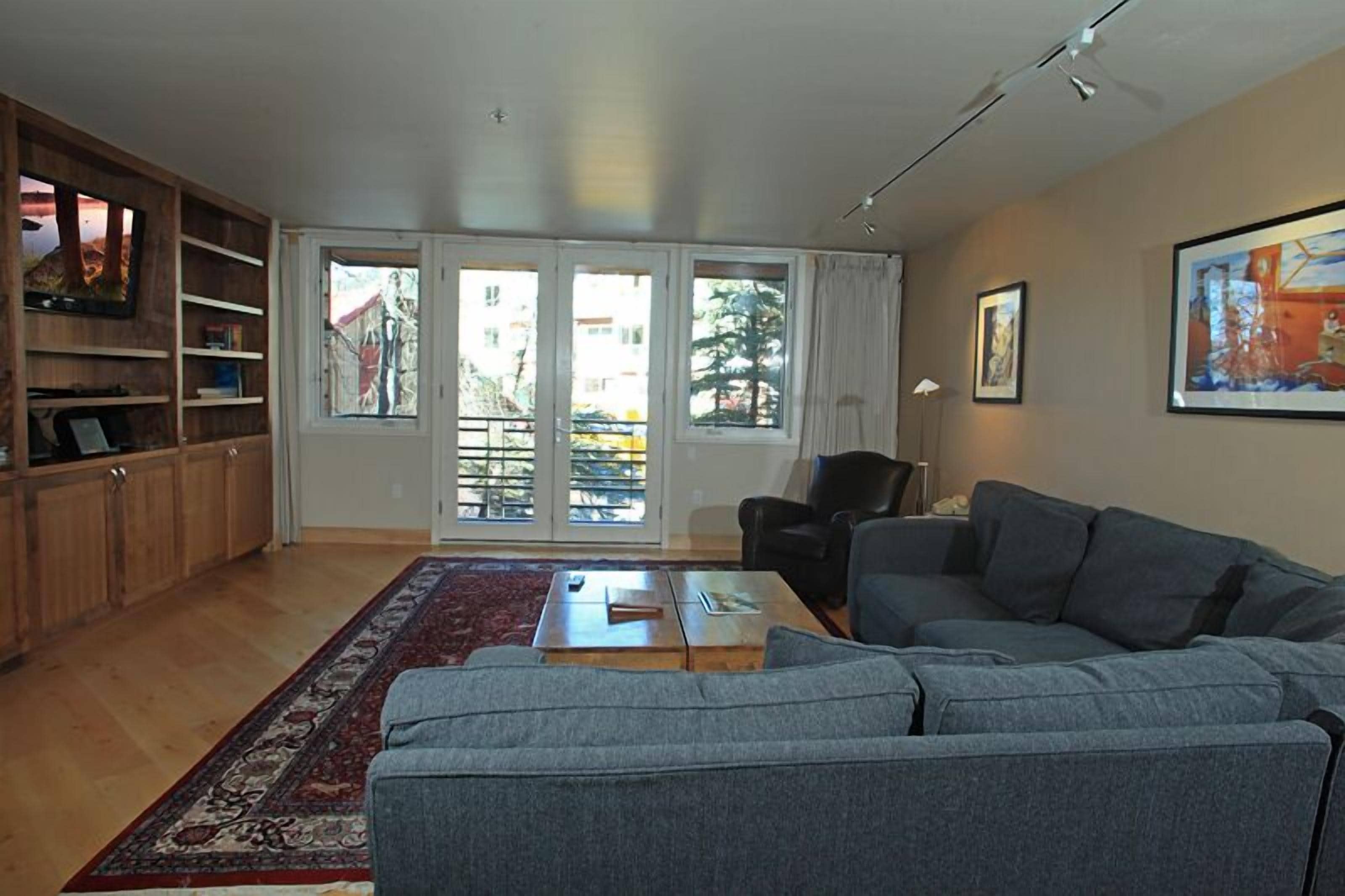 A spacious living room with a grey sofa, wooden cabinets, and a large window with a view of a balcony.