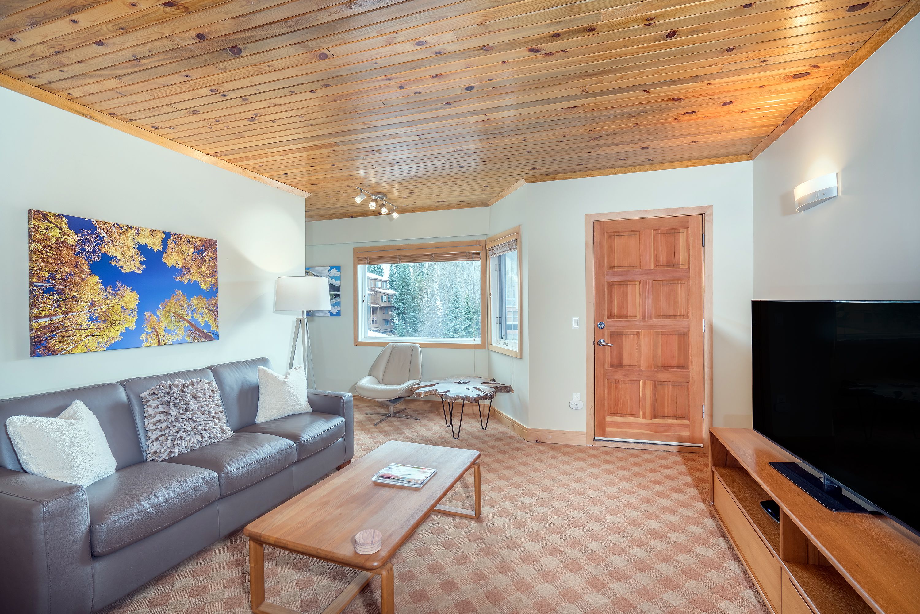 A living room with a brown leather sofa, a wooden coffee table, a flat-screen TV, and a large window offering a view of trees.
