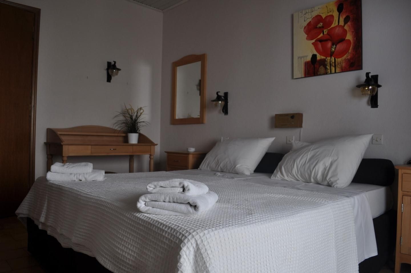 Una cama bien hecha con sábanas blancas, una mesita de noche de madera, un pequeño escritorio con una planta y un cuadro de flores rojas en la pared.