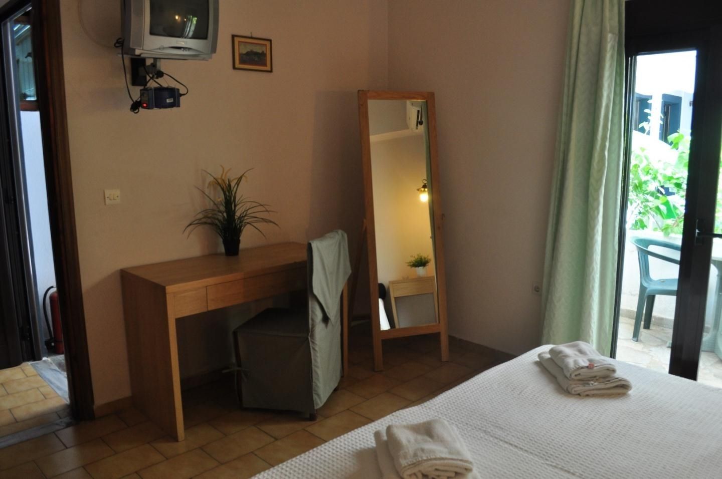 Una camera d'albergo con una scrivania in legno, una sedia, un letto con copriletto bianco, uno specchio e una pianta in vaso.