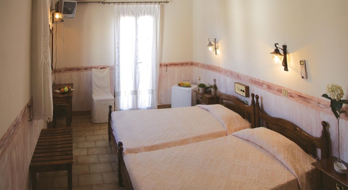 A hotel room with two beds, a small desk, a chair, a bedside table with a lamp, a window with curtains, and a wall-mounted TV.