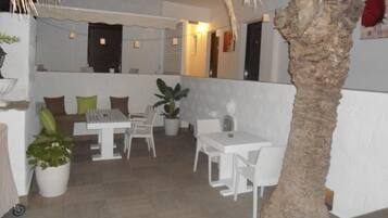 A covered outdoor seating area with white tables and chairs, potted plants, and a palm tree.
